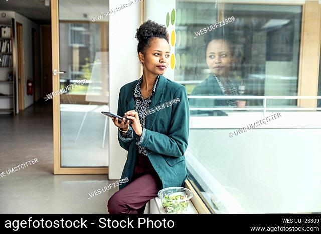 Female entrepreneur with mobile phone looking away while sitting on window sill at office
