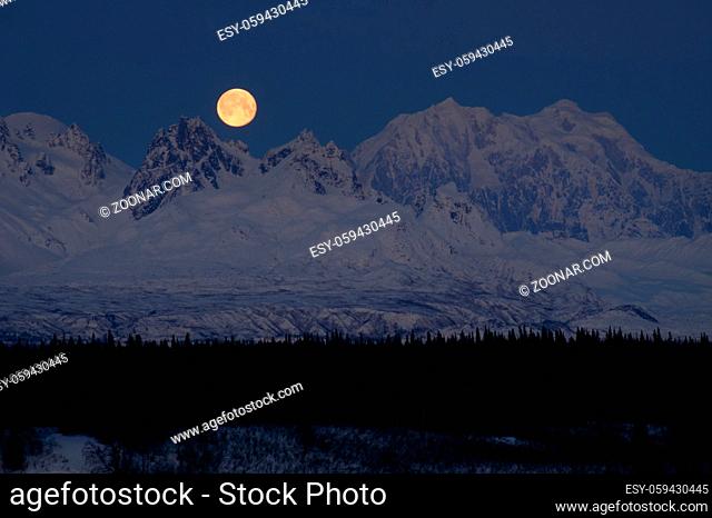 The moon emerges from behind the mountains in Denali