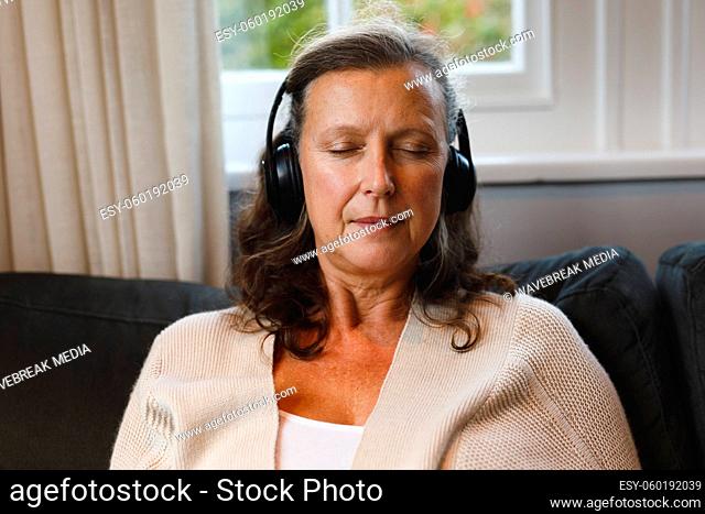 Happy senior caucasian woman in living room sitting on sofa, wearing headphones