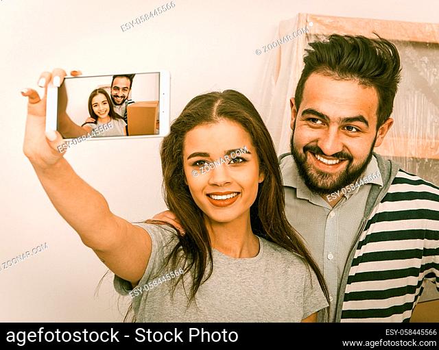 Relocation young happy couple making selfie shot in their new apartment. Man holding boxes smiling together witn young pretty woman holding phone for selfie