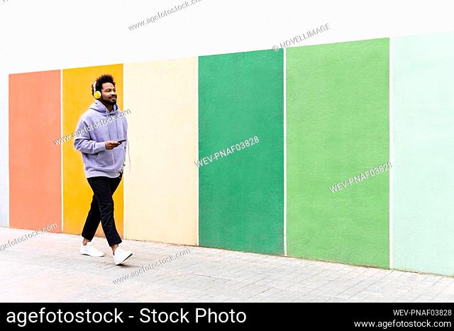 Man holding mobile phone listening music through headphones walking on footpath by colorful wall