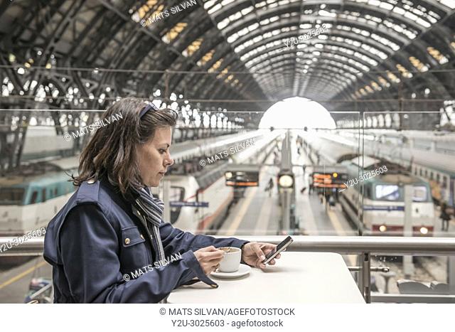 Woman Sitting in a Bar with a Cup of Coffee and Using a Smart Phone in a Train Station in Milan and Waiting in Lombardy, Italy