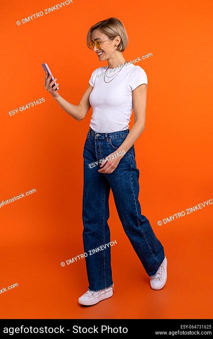 Woman blogger in white t shirt and jeans using smart phone browsing internet isolated over orange background