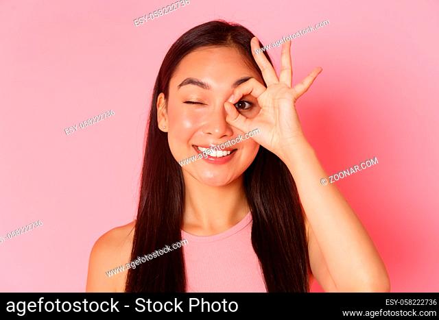 Beauty, fashion and lifestyle concept. Portrait of kawaii attractive asian girl showing okay gesture over eye and winking carefree at camera, smiling pleased