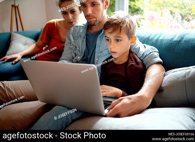 Father and son sharing laptop sitting by woman on sofa at home