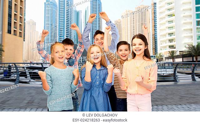 childhood, travel, tourism, gesture and people concept - happy children friends raising fists and celebrating victory over dubai city street background