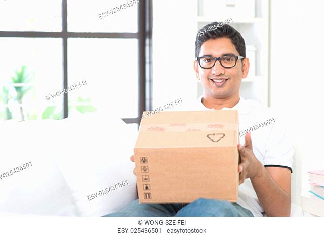 Courier delivery concept. Indian guy received an express parcel and checking the box at home. Asian man sitting on sofa indoor
