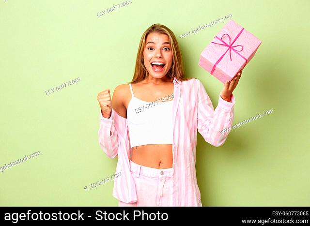 Cheerful blond girl celebrating holiday, holding gift and looking excited, standing over green background