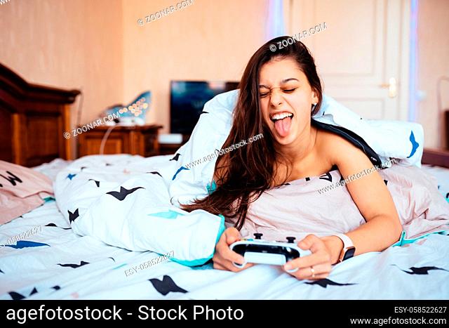 Funny girl in casual clothing lying in bed and playing video game, holding controller in hands