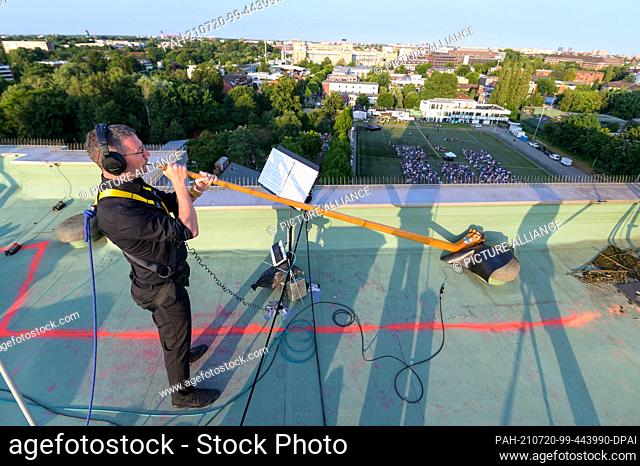 17 July 2021, Hamburg: A musician blows his alphorn on the roof of a high-rise building while hundreds of spectators listen on a football pitch and all around