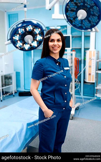 Smiling female surgeon in operating room, lamp and surgical table on background, surgery. Doctor in uniform, medical clinic worker, medicine and health
