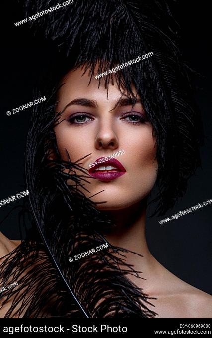 Beautiful young lady with purple make-up and big black feathers touching face. Beauty shot with glowing skin on black background. Copy space