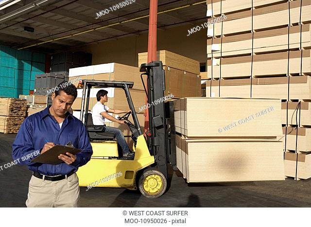 Man with clipboard in front of forklift stacking boxes