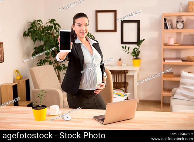 Portrait of happy and cheerful pregnant business woman smiling and demonstrating her mobile or smart phone to camera while working at home