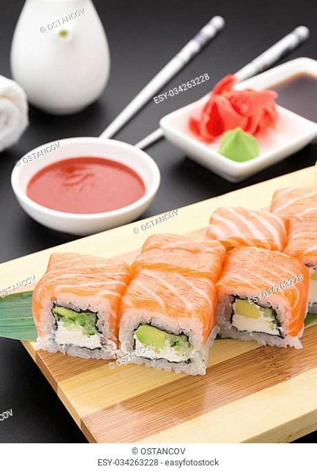 Salmon sushi roll on wooden plate ( gete ) with ginger wasabi chopsticks and sauces over black background