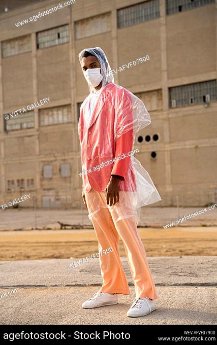Young man wearing protective face mask and raincoat standing on footpath