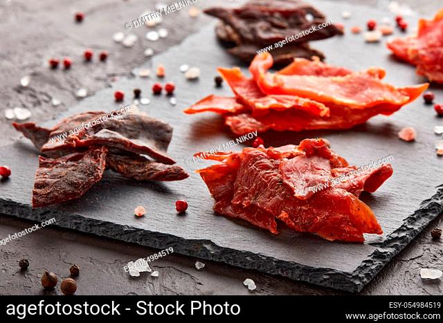 Jerky. Set of various kind of dried spiced meat on black stone cutting board on dark gray background. Snack for beer