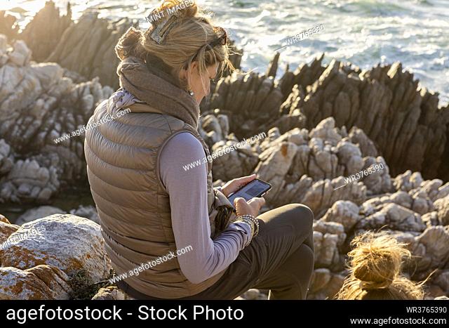 Woman sitting on rocky shore, looking at her mobile phone