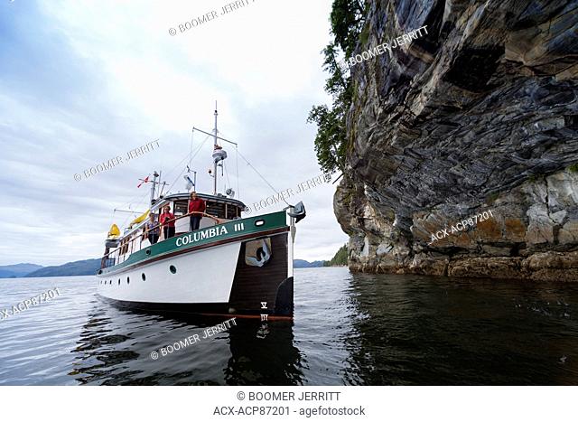 Columbia III steams in close to an overhanging rock bluff while transiting waters in the Broughton Archipelago, Central British Columbia Coast, Canada