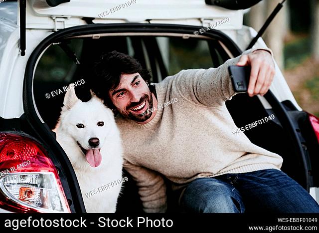 Smiling man taking selfie with dog while sitting in car trunk at forest