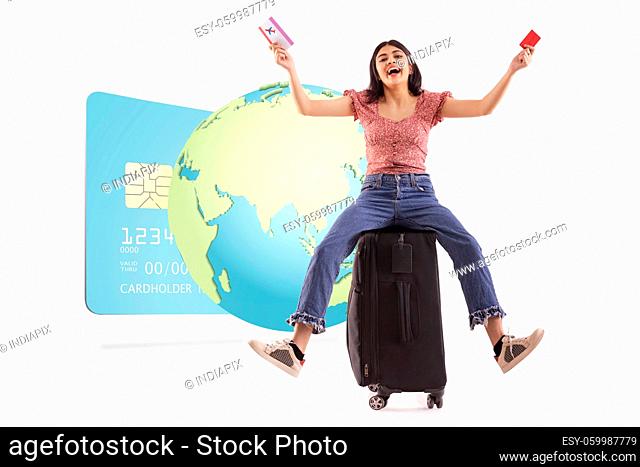 An excited woman sitting on her luggage holding credit card and ticket with a graphic of global net-banking