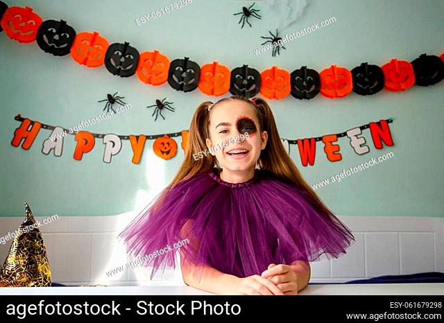 Cute little girl wearing witch hat sitting behind a table in Halloween theme decorated room. Halloween party concept. Preparation for the celebration