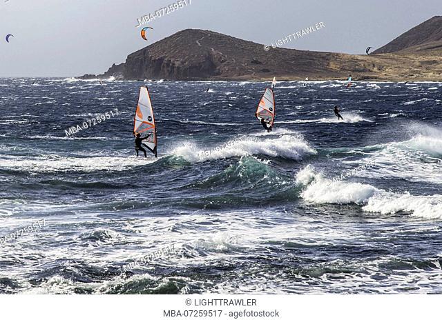 Windsurfers and kite surfers in the bay of Medano off the coast of Tenerife in strong wind. In the foreground the surf in front of the panorama of Montana Roja