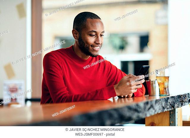 Smiling young man wearing red pullover at counter of a bar with soft drink and mobile phone