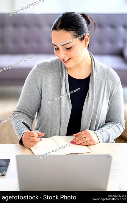 Smiling businesswoman writing in diary while sitting with laptop at home office