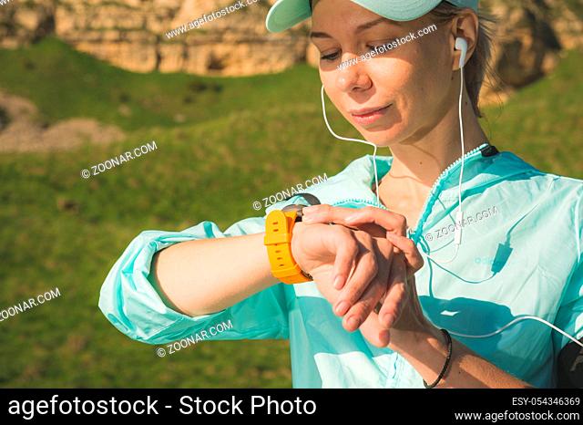 Portrait of a smiling young fitness girl in a cap and headphones checking her smart clock while sitting outdoors against a background of rocks
