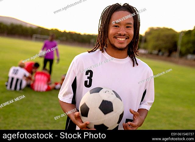 Portrait of smiling biracial male player in white uniform holding soccer ball standing at playground