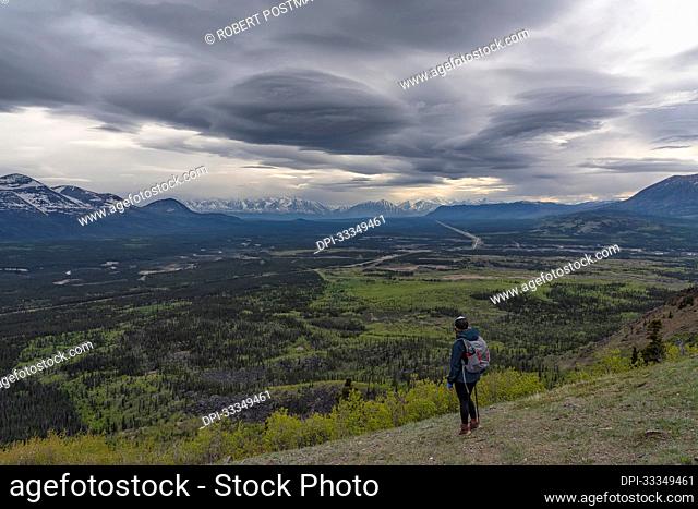 View taken from behind of a woman hiker standing on a mountainside looking out at the view of the valley and mountain ranges in the distance under a stormy sky;...