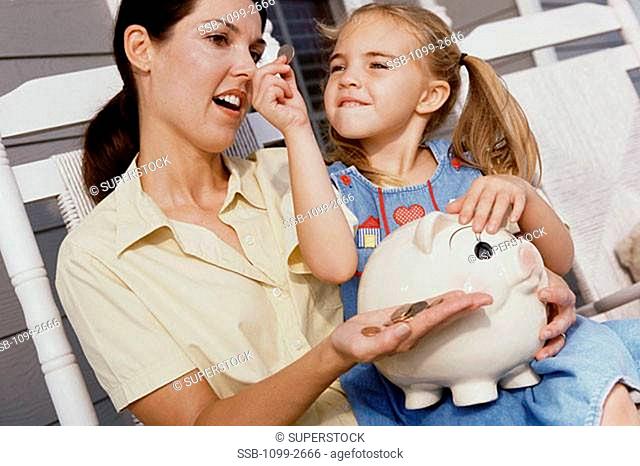 Girl sitting on her mother's lap and putting coins into a piggy bank