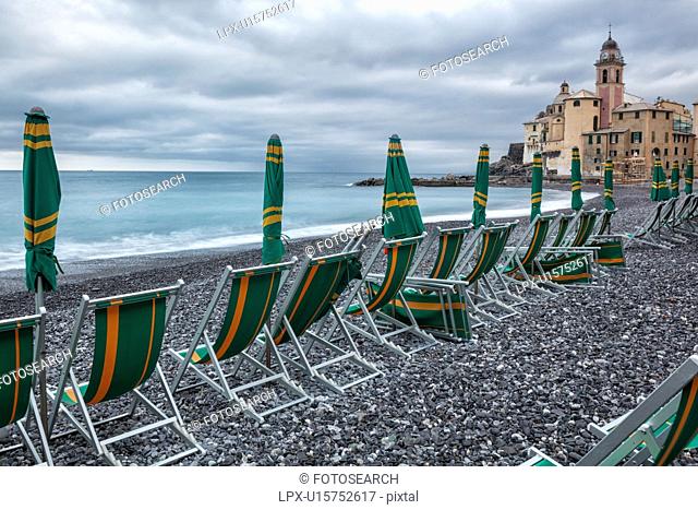 Camogli: beach and sea with waves motion blur, rows of green and yellow deck-chairs, and closed beach umbrellas, church tower and surrounding buildings beyond