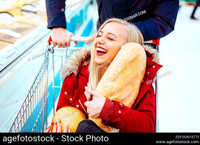 Young handsome guy rides a girl in a supermarket in a trolley. Couple have fun. Girl holds an orange and bread