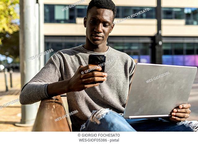 Young man sitting on a bench in the city, using laptop and smartphone