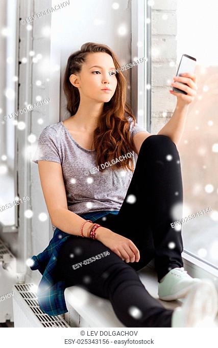 people, technology, winter, christmas and teens concept - sad unhappy pretty teenage girl sitting on windowsill with smartphone and texting over snow
