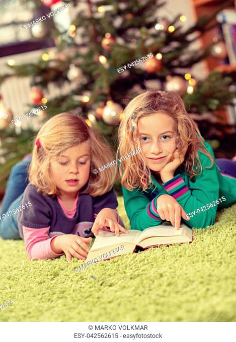 two blond girls reading a book in front of christmas tree