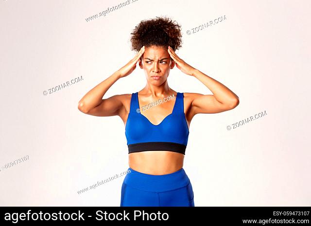 Distressed african-american female athlete in blue sport ouotfit, holding hands on head and grimacing upset, standing tensed with headache, white background
