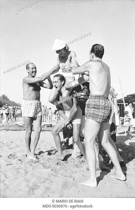 A man holding a woman on his back on the beach during the XVIII Venice International Film Festival. Venice, 1957