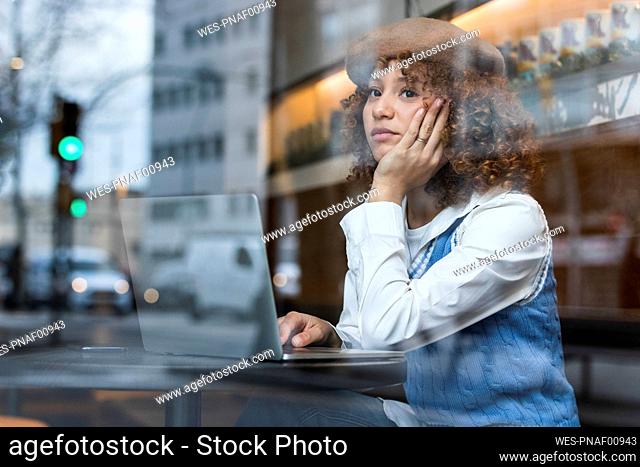 Thoughtful teenage girl sitting with hand on chin by laptop at cafe window