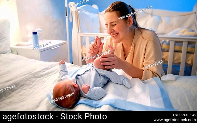 Portrait of smiling young mother smiling at her little baby son lying on bed and holding his little feet. Happy parenting and family happiness