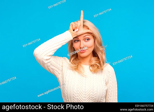 Upset unlucky woman with curly blond hair showing looser gesture holding finger near forehead, depressed with her fail. Indoor studio shot isolated on blue...