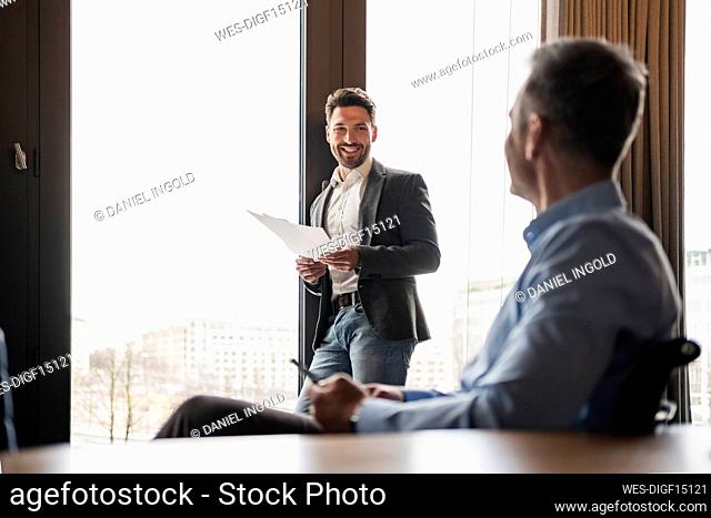 Smiling businessman with document talking to male colleague at work place