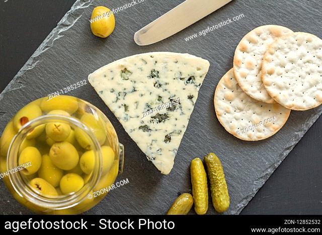 Blue cheese, green olives, pickles and crackers on black slate board - Roquefort type cheese background top view close up photo