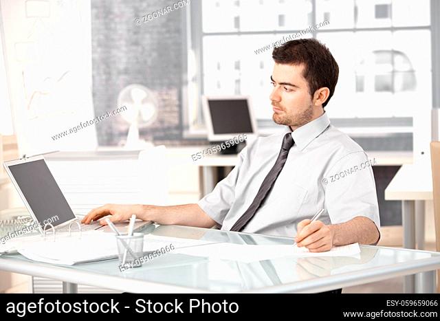 Young businessman working in bright office, using laptop, writing notes