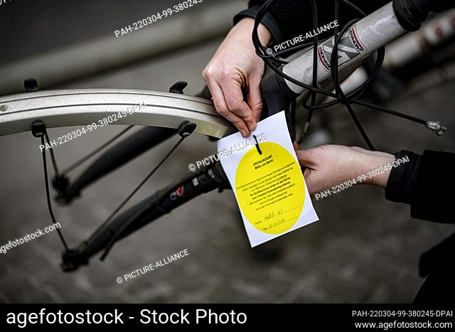 03 March 2022, Berlin: A person hangs a notice for removal by the Ordnungsamt Mitte on a scrap bicycle at the S-Bahn and U-Bahn station Wedding