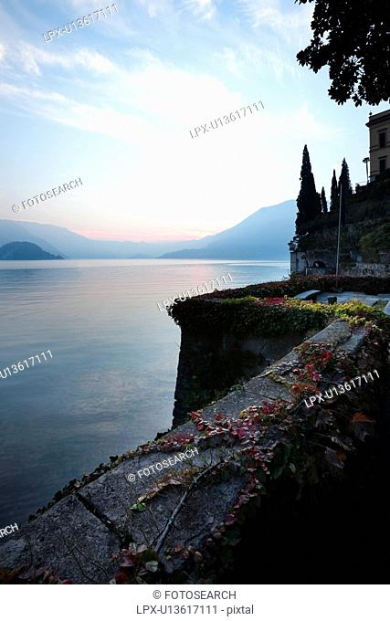 Tranquil scene overlooking Lake Como at sunset, blue and pink sky, still waters, cypress trees, wall with trailing ivy with autumn foliage, Lake Como