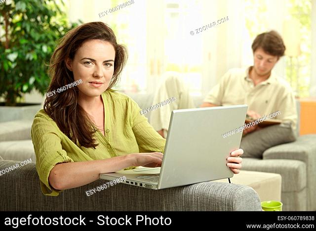 Couple at home. Woman using laptop computer, man reading book in background