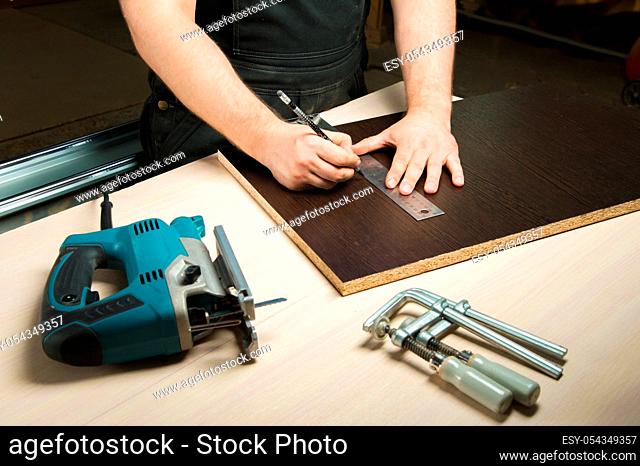 Men's hands draw a pencil on the ruler with a sub-standard template for cutting out furniture details next to the jigsaw and furniture clips